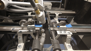 Close-up of an industrial machine with rollers, metal rods, hoses, and fasteners, likely part of a conveyor sandblast system. Components appear aligned for precise mechanical operation.