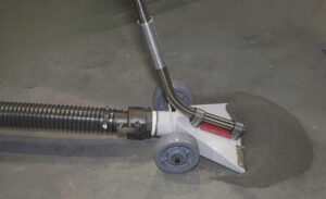 A wet vacuum cleaner nozzle with wheels, part of the PARS5000 Vacuum Recovery System, is being used to clean a liquid spill from a gray floor—ideal for maintaining cleanliness in sandblast booths.