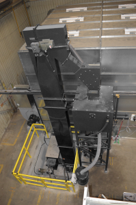 An industrial bucket elevator system with a black vertical conveyor and enclosed machinery, featuring HARS2000 Hybrid Recovery System technology, surrounded by yellow safety railings inside a large factory or warehouse setting.