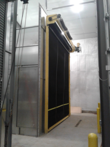 A large industrial roll-up door with black panels and yellow edges is installed in a concrete-walled Blast Booth Enclosure, featuring metal supports, white insulated walls, and a chain mechanism hanging beside the door.