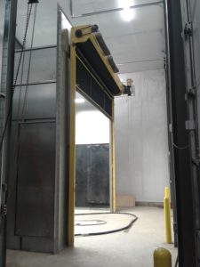 A large Blast Booth Enclosure with a yellow frame and black flexible panels is partially open in a warehouse or facility with concrete floors and white plastic walls. Cables and equipment are visible on the ground nearby.