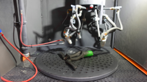 A black metal part with a green end is placed on a circular, perforated platform inside an automation-ready sandblast cabinet, surrounded by robotic arms and tubes.