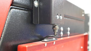 Close-up view of a machine part with black and red metal panels, bolts, and a small metallic component extending downward—designed with blasting cabinet safety in mind for secure operation.