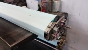 A close-up of a large industrial roller used in flexo printing, covered with a light blue sheet, partially exposing metal gears and machinery parts, set on a dark workbench in a workshop.