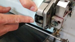 A close-up of a person's hands guiding a white material through a metal roller machine, possibly as part of an industrial process like flexo printing.