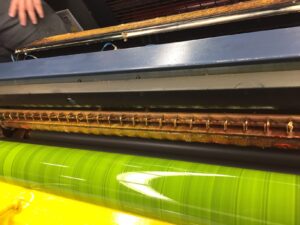 Close-up of a Flexo printing press with a bright green roller, yellow ink below, and metal components above, partially covered by a protective guard. A person’s arm is visible in the top left corner.