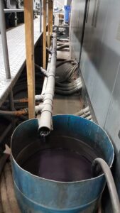 A large blue barrel filled with dark liquid sits under a long metal pipe, which is releasing liquid into the barrel—a typical scene in solvent recovery processes for litho and flexo printing. Several hoses and cables run along the industrial floor next to metal equipment.