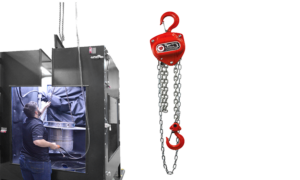 A man operates machinery inside a large industrial sandblast cabinet on the left, while a red chain hoist with hooks—ideal for parts handling solutions—hangs on the right against a plain white background.