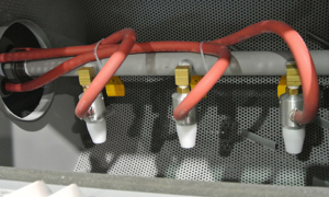 Close-up of four spray nozzles with red hoses, mounted on a metal pipe against a perforated metallic background—likely part of automation-ready sandblast cabinets or other advanced industrial equipment.