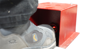 A brown shoe steps under a black, box-like device emitting a red laser beam onto the shoe, likely for measurement or alignment of ergonomic features. The background is plain white.