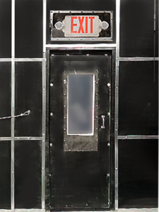 A metal door with a vertical frosted window is set in a dark wall of the blast booth, with an illuminated red EXIT sign above the door.