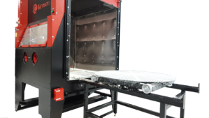 A red and black industrial machine with a window on the side, featuring a sliding metal platform with rollers and a perforated round surface in front of the blasting cabinet door.