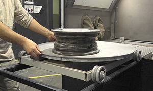 A person places a car wheel rim on a rotating platform inside a machine, possibly for cleaning or refinishing, with sandblast cabinets and parts handling solutions visible alongside tools and safety instructions.