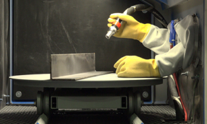 A person wearing yellow rubber gloves uses a handheld tool to work on a metal L-shaped piece inside a sandblast cabinet with access holes, highlighting efficient parts handling in a protective enclosure.