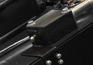 Close-up of a black metal industrial component with bolts, a cylindrical rod, and various connecting parts, possibly part of a machine or blasting cabinet door featuring essential safety devices.
