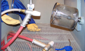 A person wearing yellow gloves and a blue protective suit works inside an automation-ready sandblast cabinet with a mesh floor, holding a hose and spray nozzle next to a perforated metal basket and a blue air blower.