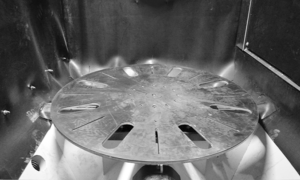 A large, circular, metallic rotating platform with evenly spaced slots is set inside an industrial Parts Handling Solutions machine, surrounded by metal walls and components.